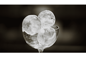 A high-contrast, black and white image of three perfectly clear, spherical Hoshizaki ice balls stacked in an elegant, stemmed cocktail glass for a premium beverage presentation