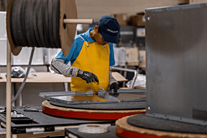 A skilled factory technician wearing a Hoshizaki cap and a protective yellow apron is using a screwdriver to assemble a metal component on the production line