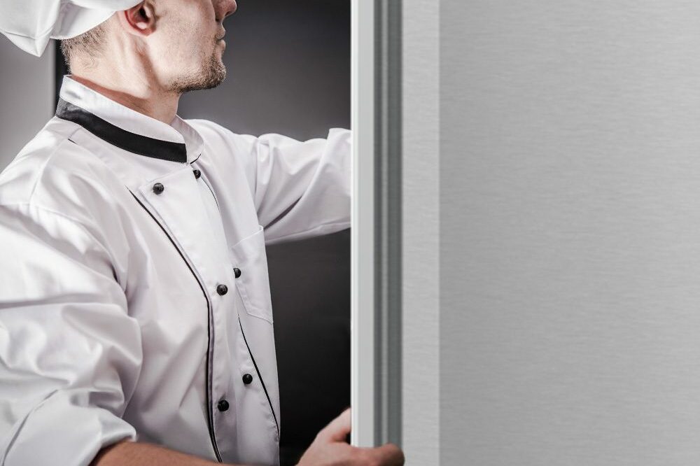 A close-up side view of a chef in a white uniform opening the seamless stainless steel door of a Hoshizaki commercial refrigerator