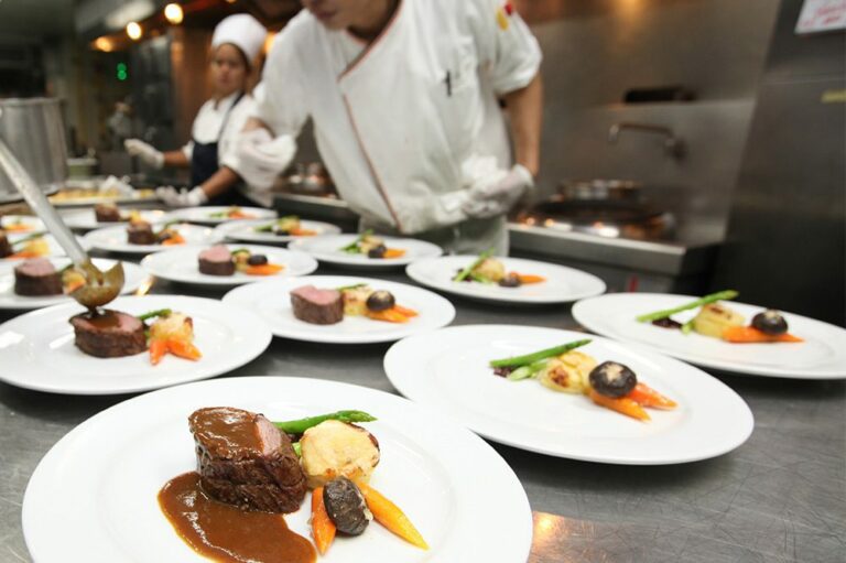 Chefs wearing white gloves are quickly plating gourmet meals at a pass-through station, relying on Hoshizaki refrigeration for ingredient freshness