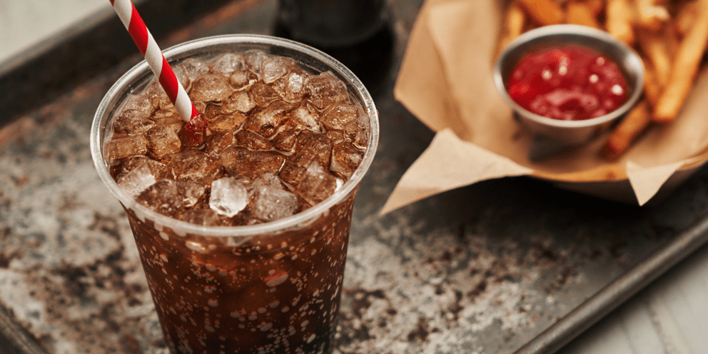 A close-up of a fizzy soda filled with Hoshizaki Cubelet chewable ice and a red and white straw, served next to an order of fries and ketchup on a metal tray
