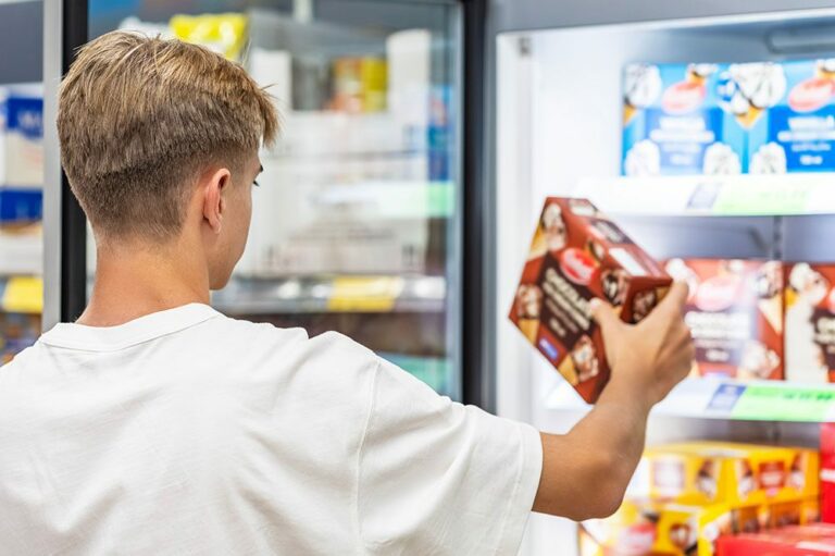 A customer holds a box of frozen treats while looking into a brightly lit Hoshizaki glass door merchandiser freezer in a retail environment