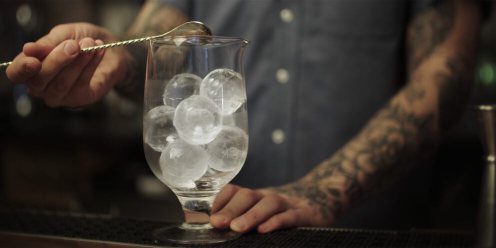 A bartender with tattooed arms stirring a cocktail in a glass filled with clear Hoshizaki Sphere Ice