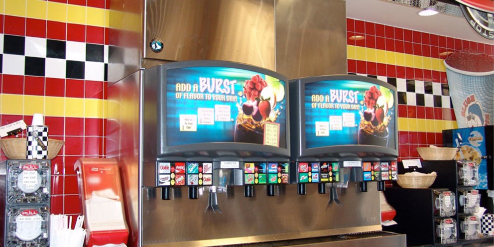 Two Hoshizaki twinpour chewable ice and beverage dispensers are featured against a vibrant yellow, red, and black tiled wall in a quick-service restaurant or convenience store